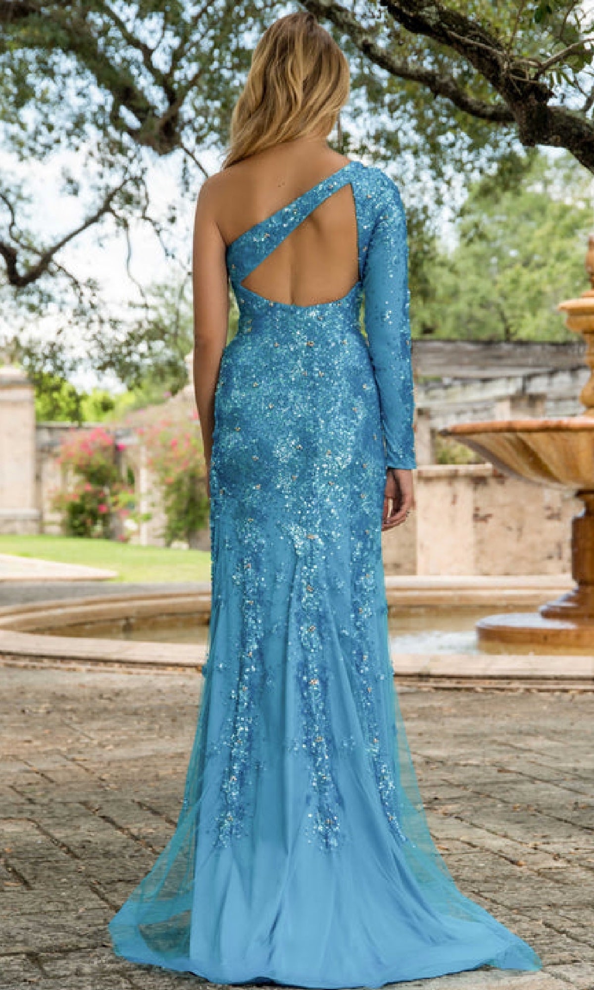 Outdoors, a woman wears the Ava Presley 28578—a striking, fitted blue sequin prom dress with a single long sleeve, shoulder drape, open back, and elegant one-shoulder design.